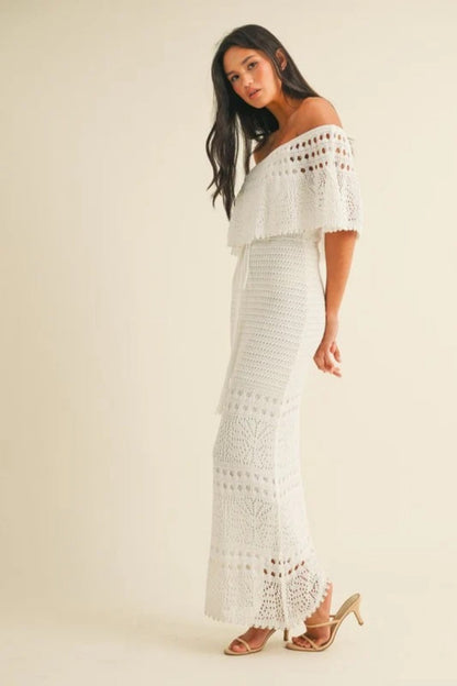 White - A woman wearing a white off-the-shoulder crochet maxi dress with a flounce bodice, a drawstring waist, and intricate open-knit patterns throughout.
