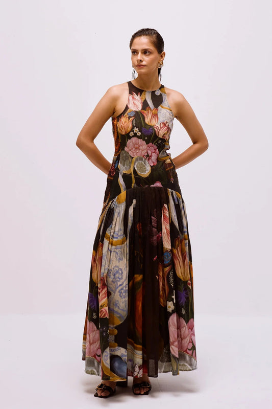 A woman with dark hair slicked back stands against a white background, wearing a full-length, sleeveless halter dress. The dress features a dark, intricate print of flowers, lemons, and antique vases, with a fitted bodice and a long, pleated A-line skirt.