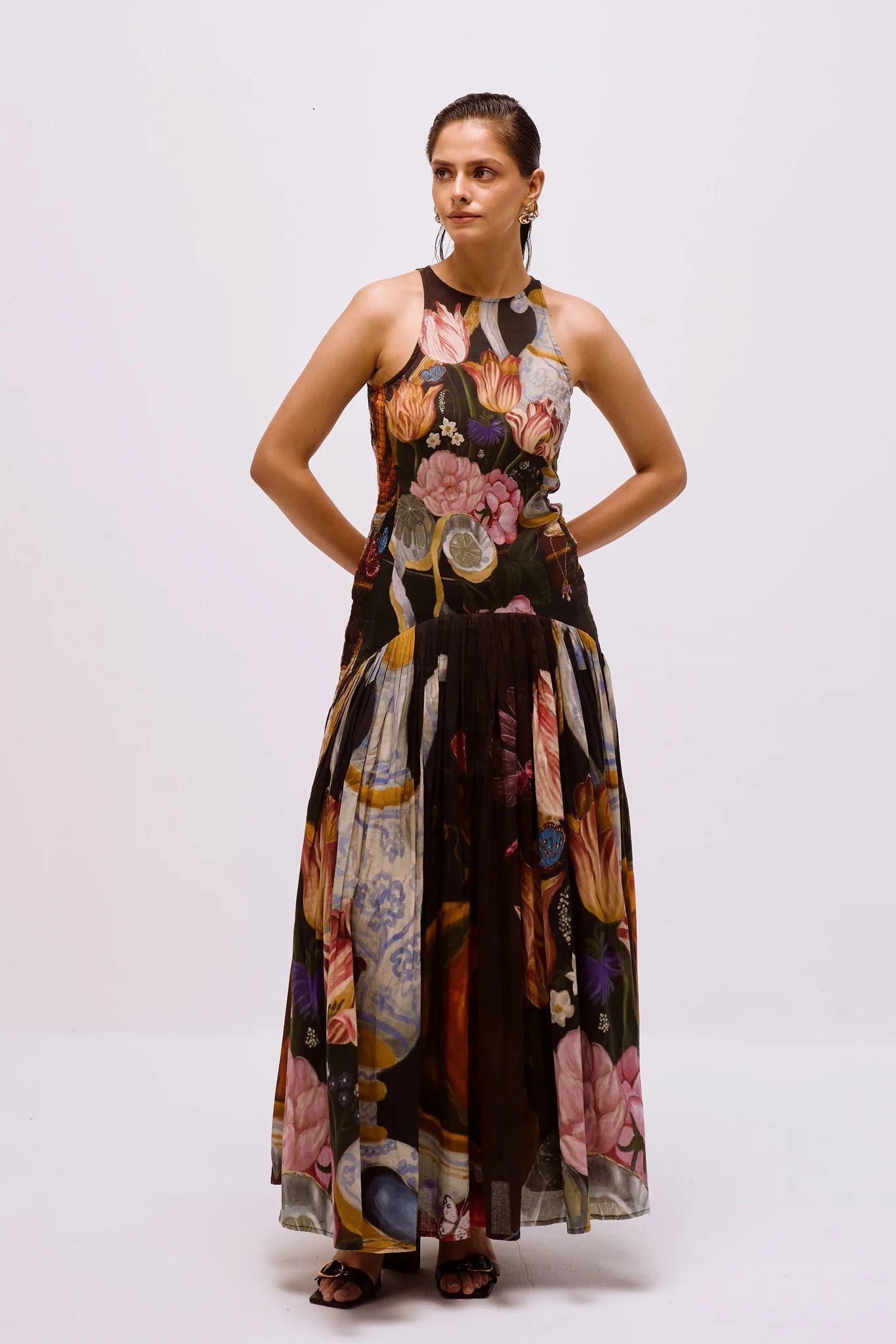 A woman with dark hair pulled back stands against a white background, wearing a sleeveless, high-neck maxi dress featuring a dark, painterly still-life print of flowers and vases.