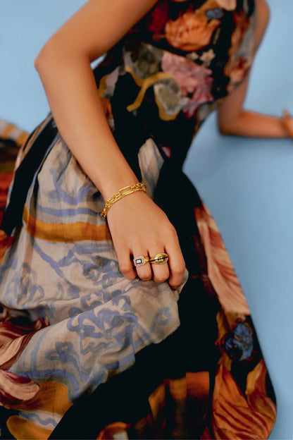 A woman with dark hair slicked back stands against a white background, wearing a full-length, sleeveless halter dress. The dress features a dark, intricate print of flowers, lemons, and antique vases, with a fitted bodice and a long, pleated A-line skirt.