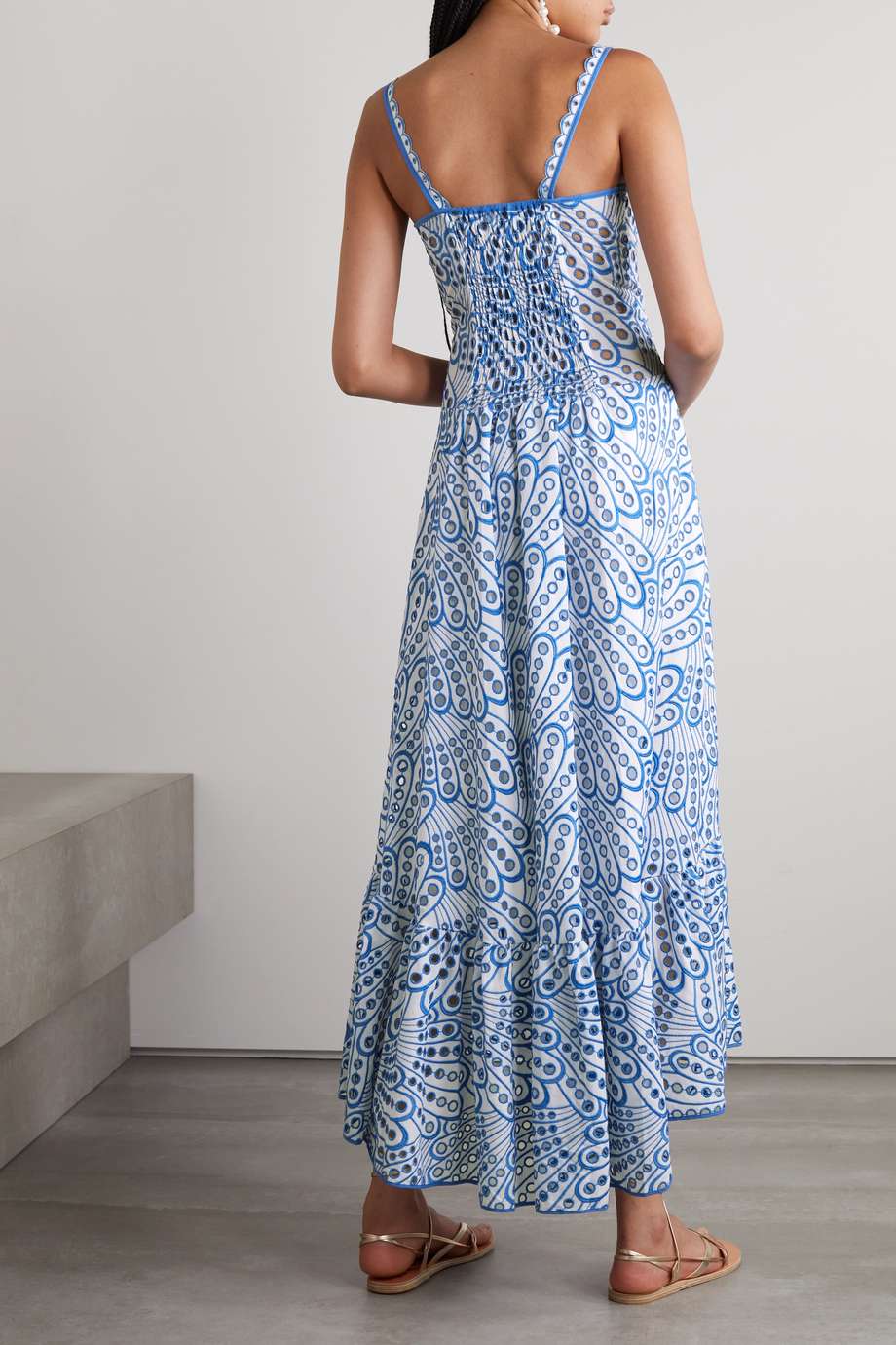 A woman models a sleeveless blue and white geometric print midi dress with a square neckline, tiered skirt, and eyelet cutouts, standing against a white wall. She pairs the dress with gold lace-up flat sandals and a woven tote bag.