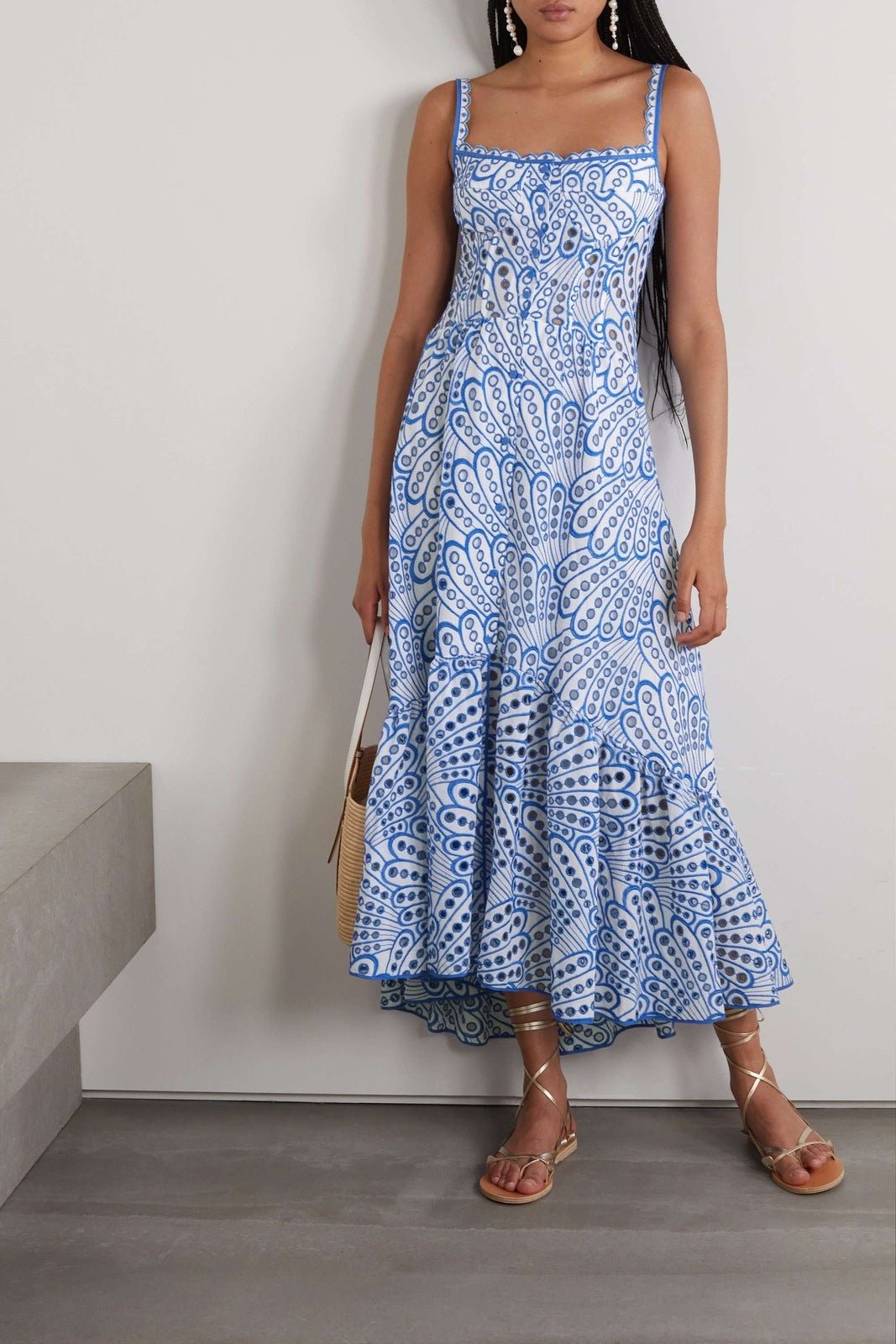 A woman models a sleeveless blue and white geometric print midi dress with a square neckline, tiered skirt, and eyelet cutouts, standing against a white wall. She pairs the dress with gold lace-up flat sandals and a woven tote bag.