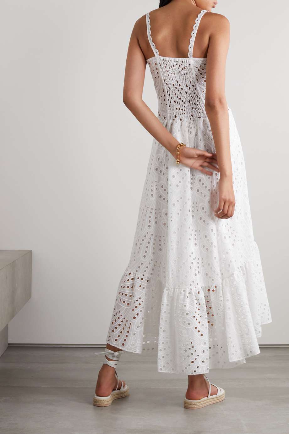 A woman models a sleeveless blue and white geometric print midi dress with a square neckline, tiered skirt, and eyelet cutouts, standing against a white wall. She pairs the dress with gold lace-up flat sandals and a woven tote bag.