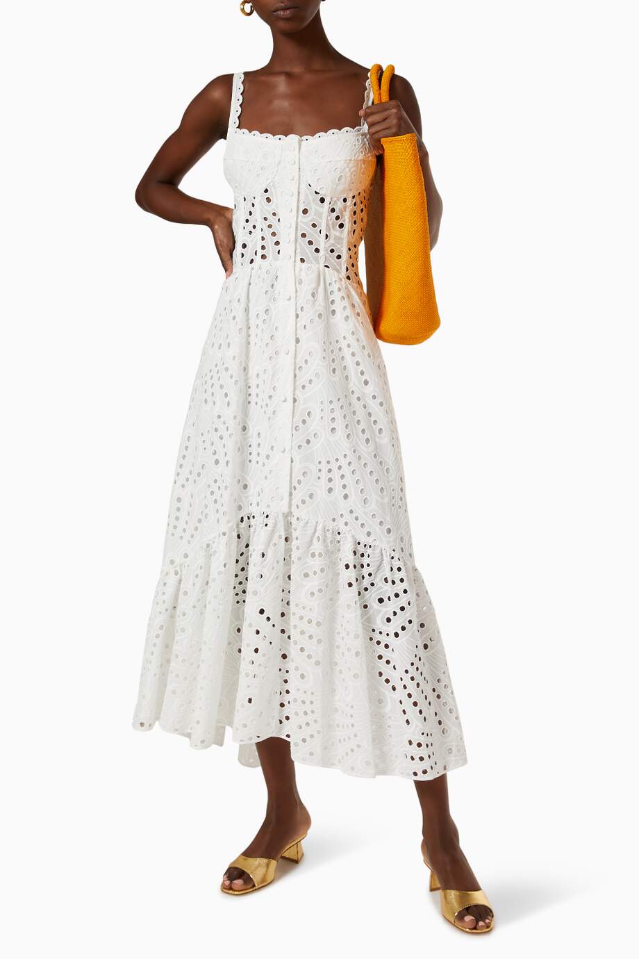 A woman models a sleeveless blue and white geometric print midi dress with a square neckline, tiered skirt, and eyelet cutouts, standing against a white wall. She pairs the dress with gold lace-up flat sandals and a woven tote bag.