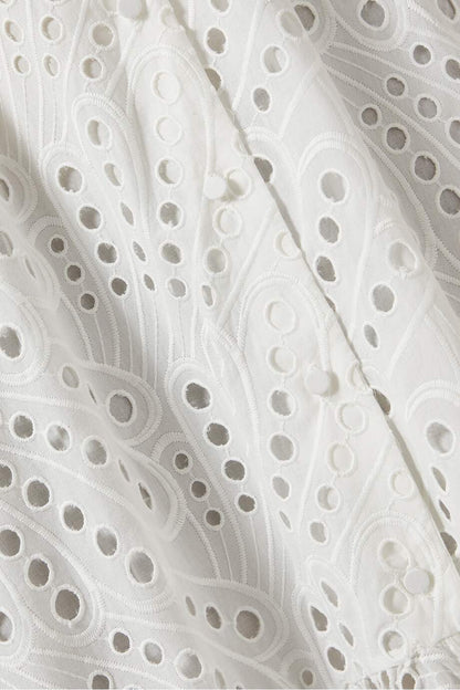 A woman models a sleeveless blue and white geometric print midi dress with a square neckline, tiered skirt, and eyelet cutouts, standing against a white wall. She pairs the dress with gold lace-up flat sandals and a woven tote bag.