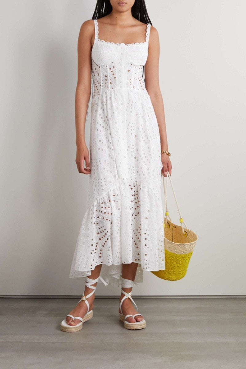 A woman models a sleeveless blue and white geometric print midi dress with a square neckline, tiered skirt, and eyelet cutouts, standing against a white wall. She pairs the dress with gold lace-up flat sandals and a woven tote bag.