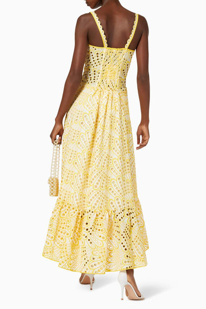 A woman models a sleeveless blue and white geometric print midi dress with a square neckline, tiered skirt, and eyelet cutouts, standing against a white wall. She pairs the dress with gold lace-up flat sandals and a woven tote bag.