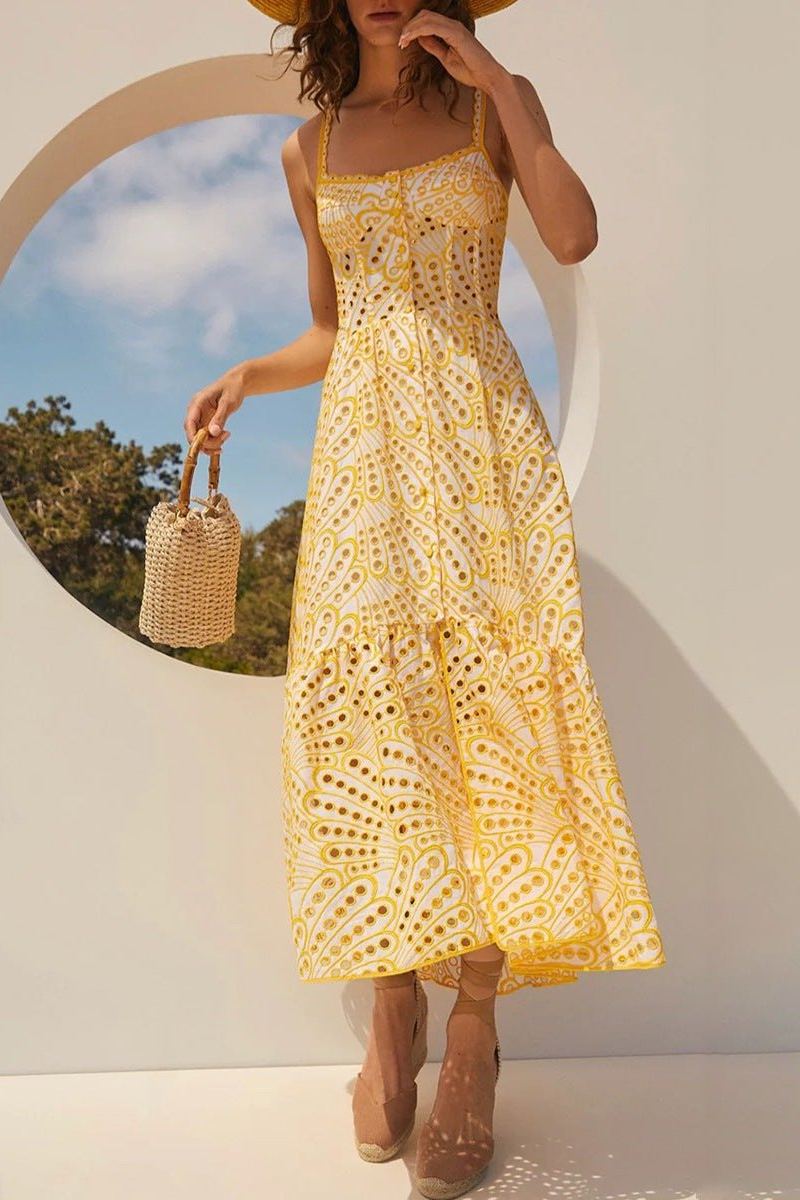 A woman models a sleeveless blue and white geometric print midi dress with a square neckline, tiered skirt, and eyelet cutouts, standing against a white wall. She pairs the dress with gold lace-up flat sandals and a woven tote bag.