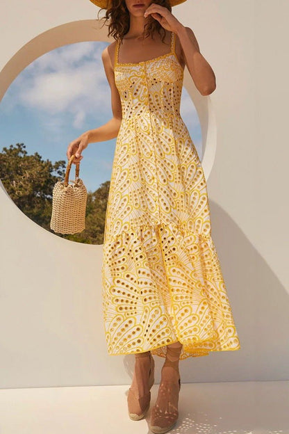 A woman models a sleeveless blue and white geometric print midi dress with a square neckline, tiered skirt, and eyelet cutouts, standing against a white wall. She pairs the dress with gold lace-up flat sandals and a woven tote bag.