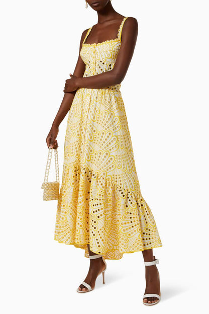 A woman models a sleeveless blue and white geometric print midi dress with a square neckline, tiered skirt, and eyelet cutouts, standing against a white wall. She pairs the dress with gold lace-up flat sandals and a woven tote bag.