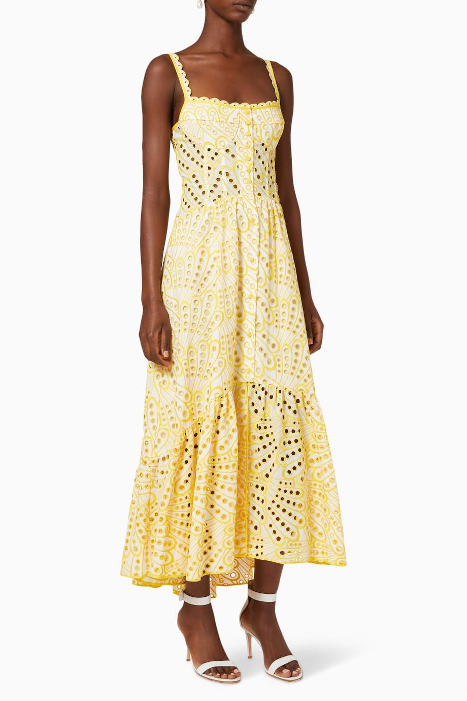A woman models a sleeveless blue and white geometric print midi dress with a square neckline, tiered skirt, and eyelet cutouts, standing against a white wall. She pairs the dress with gold lace-up flat sandals and a woven tote bag.