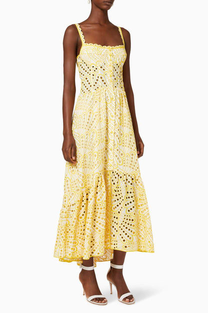 A woman models a sleeveless blue and white geometric print midi dress with a square neckline, tiered skirt, and eyelet cutouts, standing against a white wall. She pairs the dress with gold lace-up flat sandals and a woven tote bag.