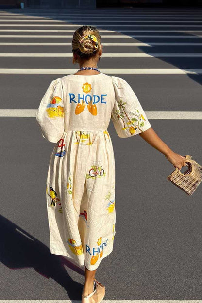 A woman wears a cream-colored, v-neck midi dress with elbow-length sleeves and a tropical vacation print. She holds a large brown woven tote bag and stands against a plain white background.