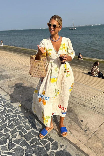 A woman wears a cream-colored, v-neck midi dress with elbow-length sleeves and a tropical vacation print. She holds a large brown woven tote bag and stands against a plain white background.
