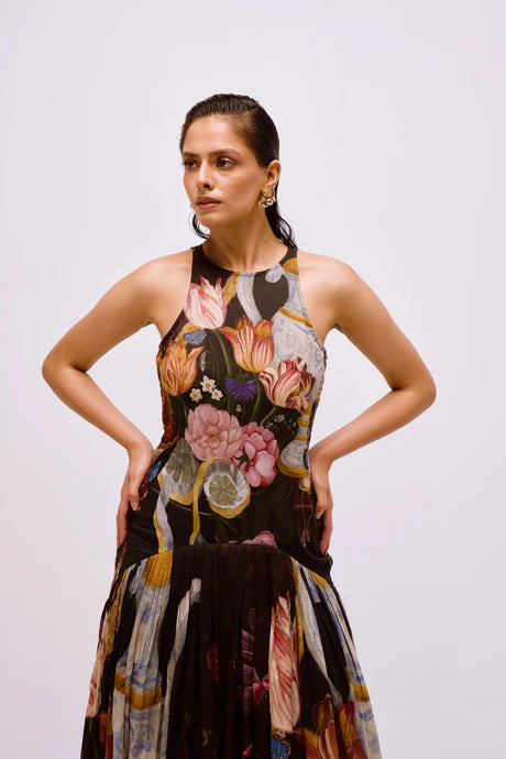 A woman with dark hair pulled back stands against a white background, wearing a sleeveless, high-neck maxi dress featuring a dark, painterly still-life print of flowers and vases.