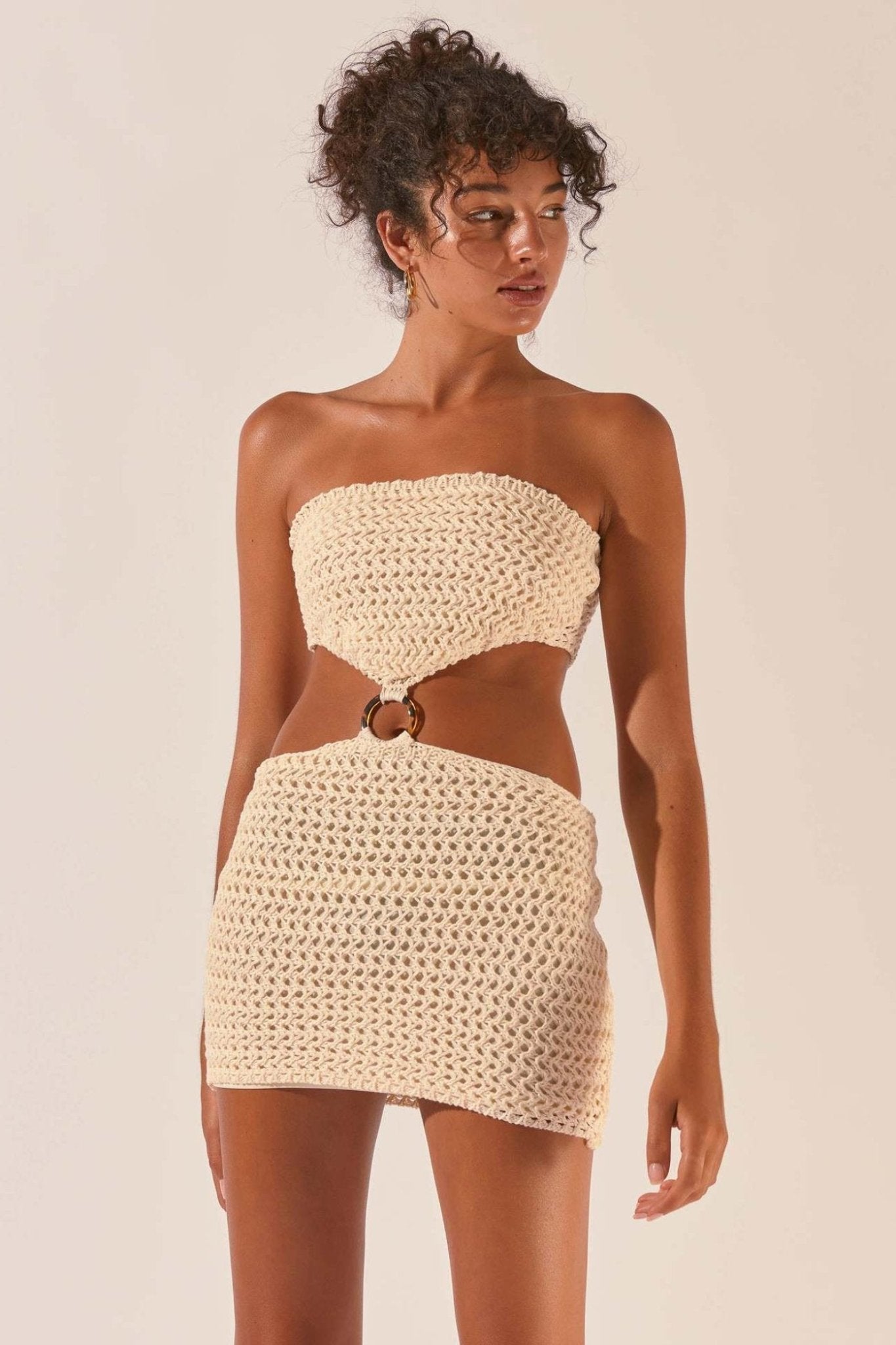 A woman with her hair up models a cream-colored, strapless crochet mini dress. The dress has a large cutout at the waist, with the top and bottom sections connected by a brown, tortoiseshell-patterned O-ring.