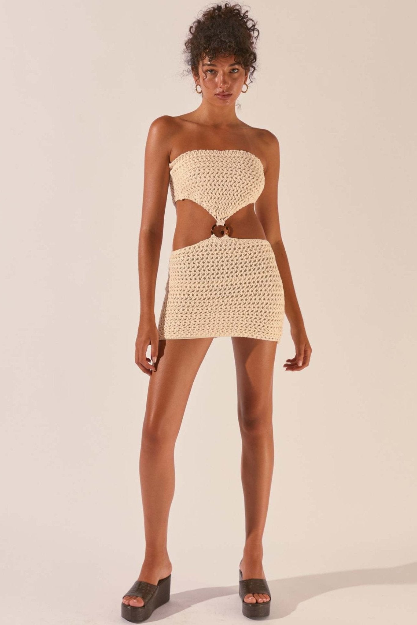 A woman wearing a cream-colored crochet knit mini dress featuring a strapless top, a fitted skirt, and a ring detail connecting the two pieces at the midriff.