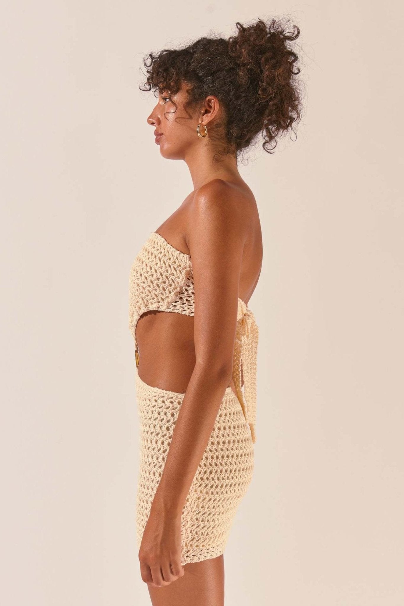 A woman wearing a cream-colored crochet knit mini dress featuring a strapless top, a fitted skirt, and a ring detail connecting the two pieces at the midriff.