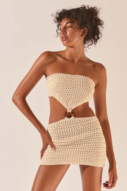 A woman wearing a cream-colored crochet knit mini dress featuring a strapless top, a fitted skirt, and a ring detail connecting the two pieces at the midriff.
