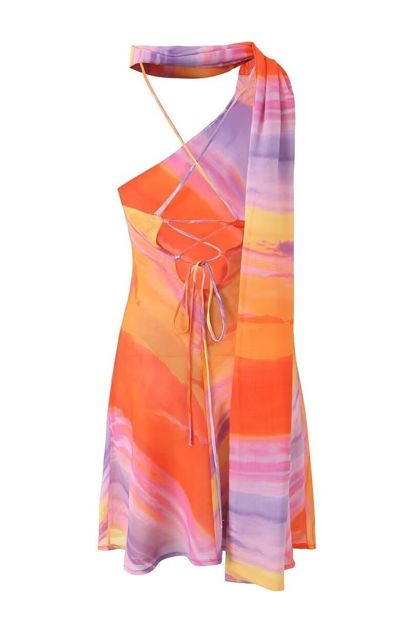 A woman wearing a one-shoulder watercolor print mini dress with a halter neck, standing outdoors in front of a colorful building and palm trees.