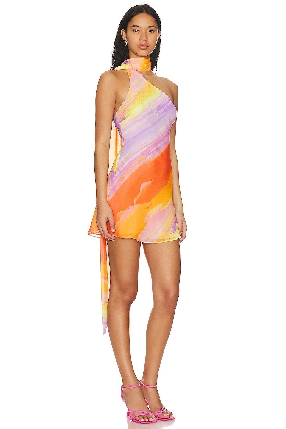 A woman wearing a one-shoulder watercolor print mini dress with a halter neck, standing outdoors in front of a colorful building and palm trees.