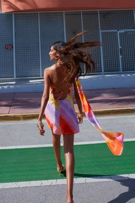 A woman wearing a one-shoulder watercolor print mini dress with a halter neck, standing outdoors in front of a colorful building and palm trees.