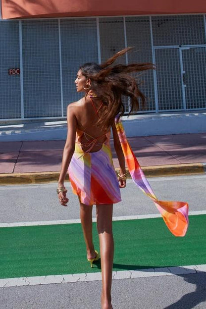 A woman wearing a one-shoulder watercolor print mini dress with a halter neck, standing outdoors in front of a colorful building and palm trees.