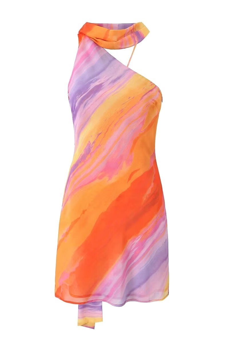 A woman wearing a one-shoulder watercolor print mini dress with a halter neck, standing outdoors in front of a colorful building and palm trees.