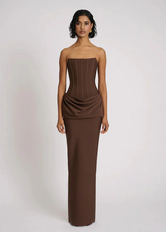 A woman wearing a formal, floor-length brown strapless dress featuring a structured corset bodice with vertical seaming and an elegant draped fabric detail at the hips.