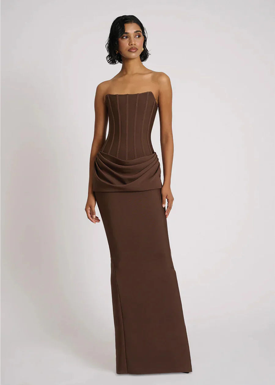 A woman wearing a formal, floor-length brown strapless dress featuring a structured corset bodice with vertical seaming and an elegant draped fabric detail at the hips.
