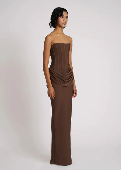 A woman wearing a formal, floor-length brown strapless dress featuring a structured corset bodice with vertical seaming and an elegant draped fabric detail at the hips.