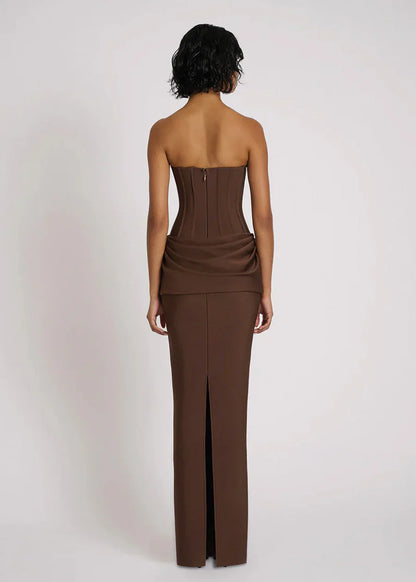 A woman wearing a formal, floor-length brown strapless dress featuring a structured corset bodice with vertical seaming and an elegant draped fabric detail at the hips.