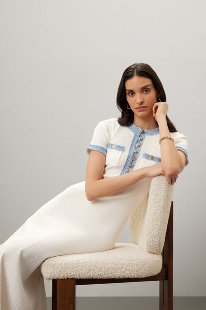 A woman models a short-sleeved, cream-colored ribbed knit midi dress with a button placket and contrasting light blue frayed trim on the neckline, pockets, and sleeves. She is holding a small grey handbag and wearing dark blue ballet flats against a plain grey background.