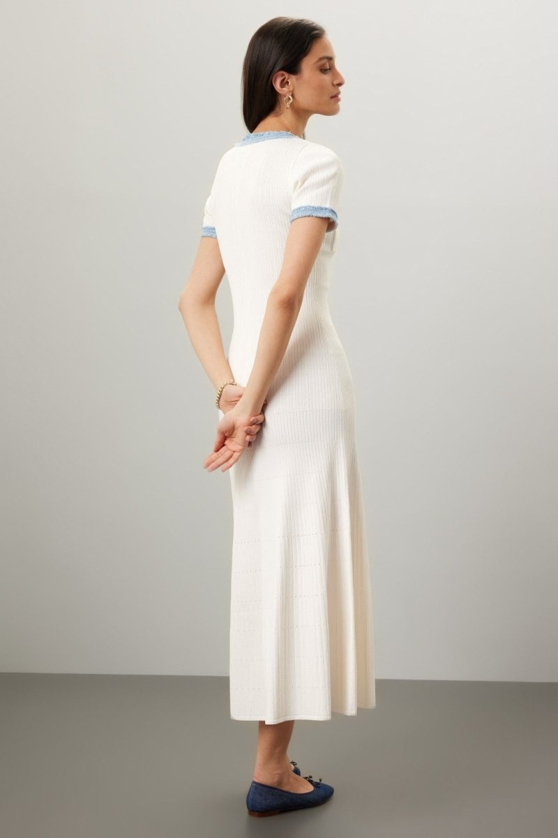 A woman models a short-sleeved, cream-colored ribbed knit midi dress with a button placket and contrasting light blue frayed trim on the neckline, pockets, and sleeves. She is holding a small grey handbag and wearing dark blue ballet flats against a plain grey background.