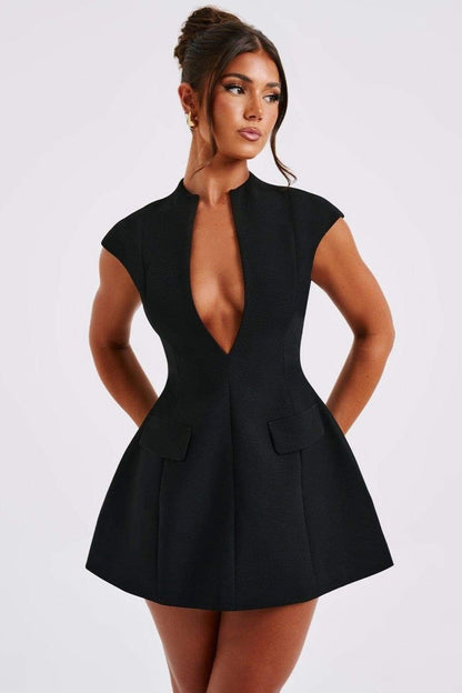 A woman models a black structured mini blazer dress featuring a high mock neck, a deep plunging keyhole cutout, extended cap sleeves, and a flared A-line skirt with flap pockets.