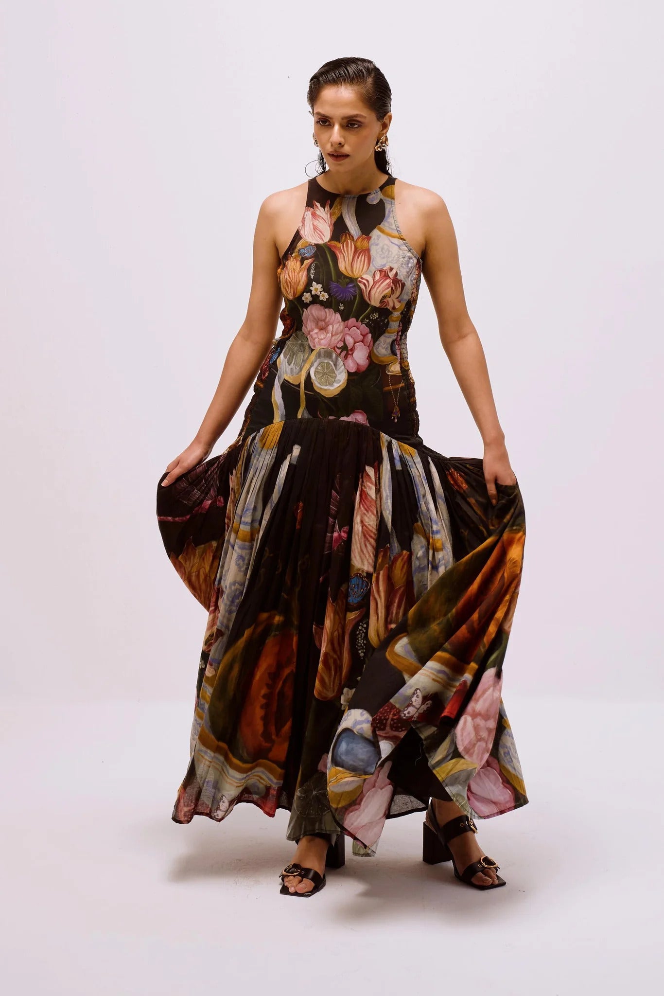 A woman with dark hair slicked back stands against a white background, wearing a full-length, sleeveless halter dress. The dress features a dark, intricate print of flowers, lemons, and antique vases, with a fitted bodice and a long, pleated A-line skirt.