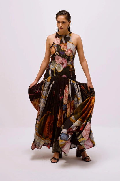 A woman with dark hair slicked back stands against a white background, wearing a full-length, sleeveless halter dress. The dress features a dark, intricate print of flowers, lemons, and antique vases, with a fitted bodice and a long, pleated A-line skirt.
