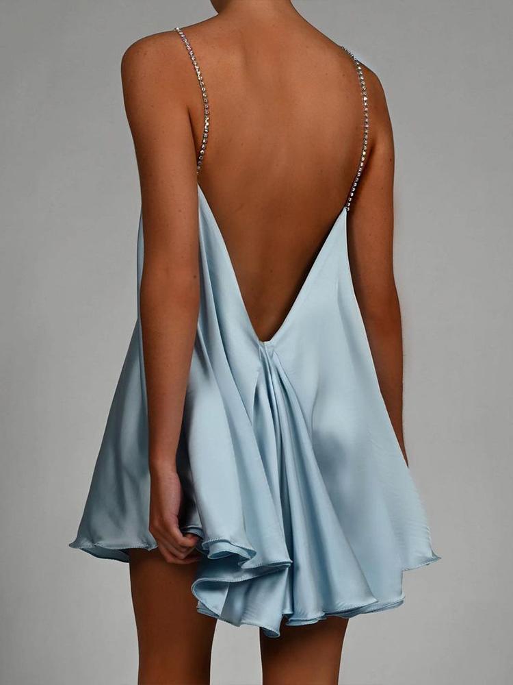 A woman wearing a chartreuse green satin mini dress with a plunging V-neckline created by a sparkling crystal chain harness that forms the straps and crosses over the chest. The dress has a loose, A-line silhouette and a ruffled hem, shown in motion against a grey background.