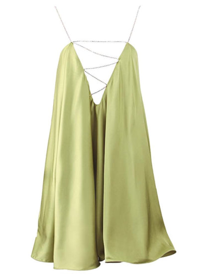 A woman wearing a chartreuse green satin mini dress with a plunging V-neckline created by a sparkling crystal chain harness that forms the straps and crosses over the chest. The dress has a loose, A-line silhouette and a ruffled hem, shown in motion against a grey background.