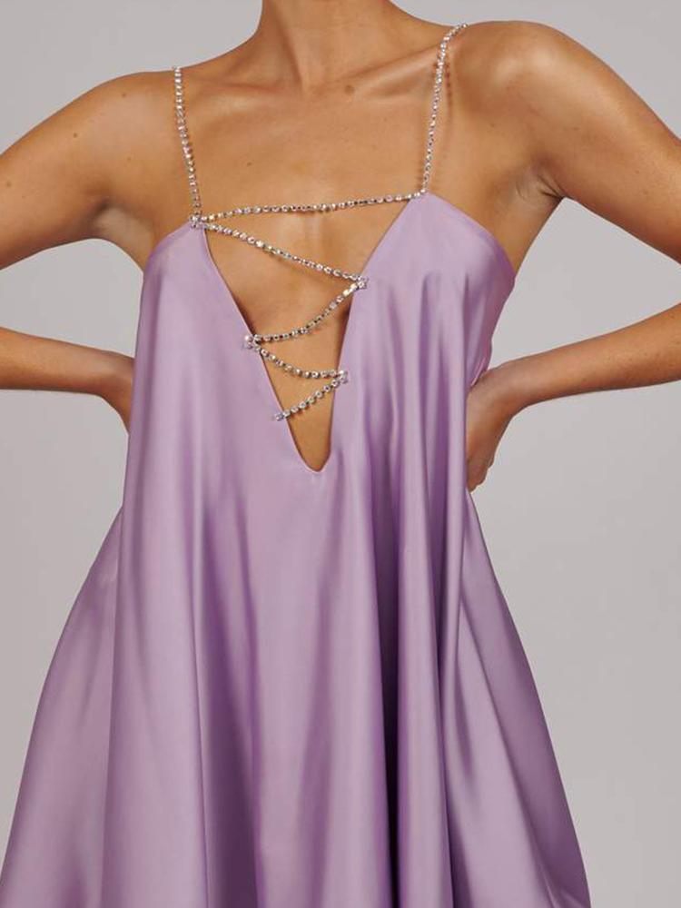 A woman wearing a chartreuse green satin mini dress with a plunging V-neckline created by a sparkling crystal chain harness that forms the straps and crosses over the chest. The dress has a loose, A-line silhouette and a ruffled hem, shown in motion against a grey background.
