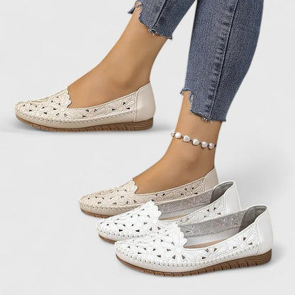 Women's slip-on loafers featuring a faux-leather upper with intricate floral cutouts and a flat, textured sole.