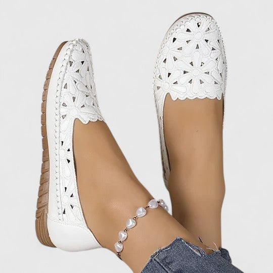 Women's slip-on loafers featuring a faux-leather upper with intricate floral cutouts and a flat, textured sole.
