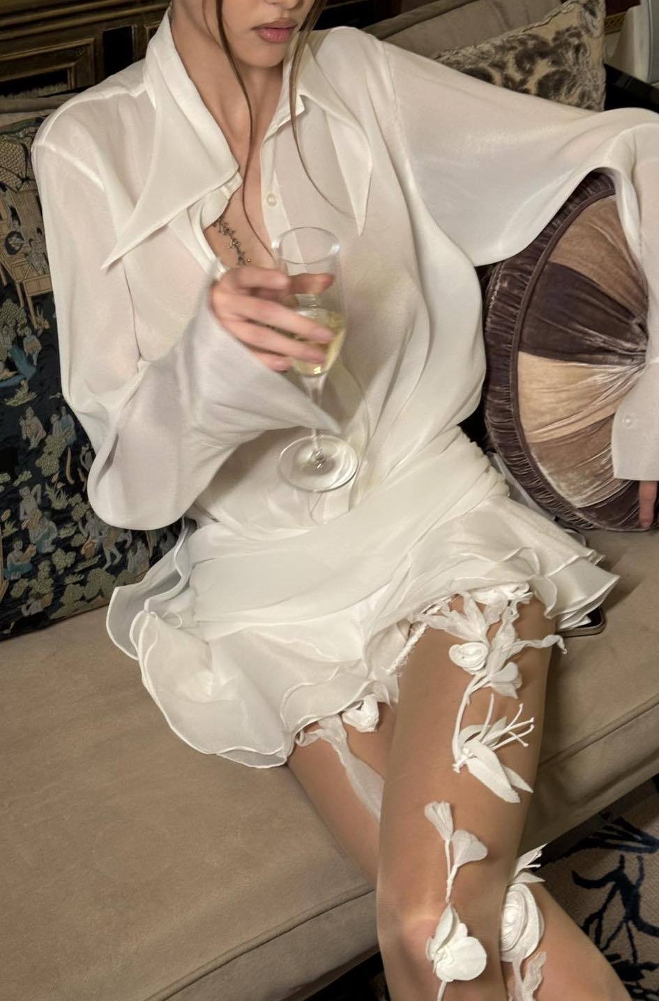 Front view of a woman wearing a sheer white mini shirt dress featuring a pointed collar, blouson top, wide ruched waist, and a short, tiered ruffled skirt.