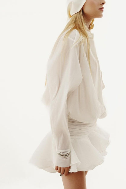 A model wearing a semi-sheer white long-sleeve shirt dress featuring a draped blouson top, a wide ruched waist, and a layered ruffled mini skirt.