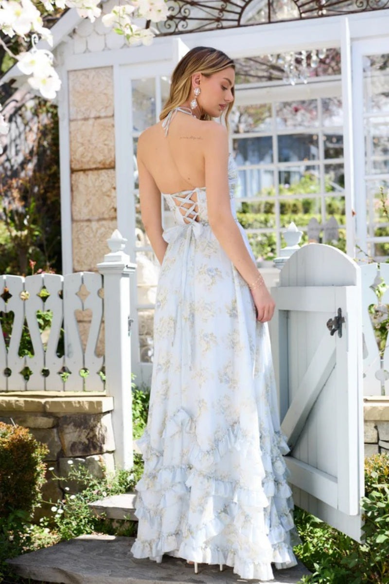Blue - A woman posing in a light blue floral chiffon maxi dress with a beaded halter neckline, a high thigh-high slit, and a tiered ruffled hem, standing in front of a white garden gate.