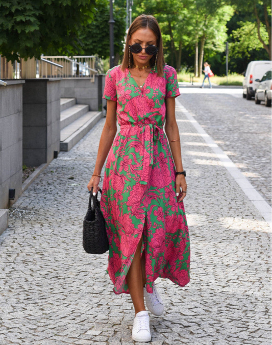 Dark Pink - A woman walking on a cobblestone street wearing a vibrant pink and green floral print midi dress with a V-neckline, short sleeves, a tie-waist, and a front slit.