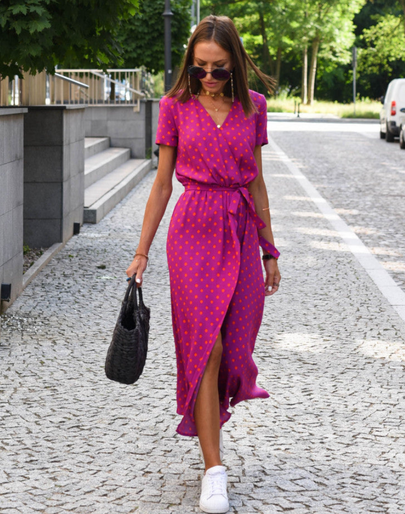 Fuschia - A woman walking on a cobblestone street wearing a vibrant pink and green floral print midi dress with a V-neckline, short sleeves, a tie-waist, and a front slit.
