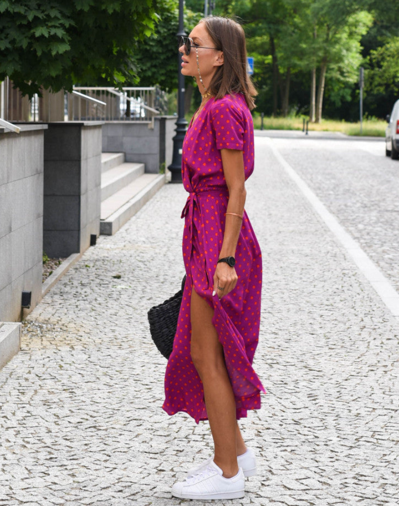Fuschia - A woman walking on a cobblestone street wearing a vibrant pink and green floral print midi dress with a V-neckline, short sleeves, a tie-waist, and a front slit.