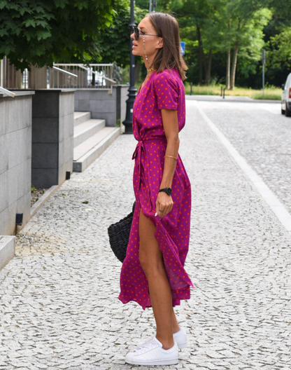 Fuschia - A woman walking on a cobblestone street wearing a vibrant pink and green floral print midi dress with a V-neckline, short sleeves, a tie-waist, and a front slit.