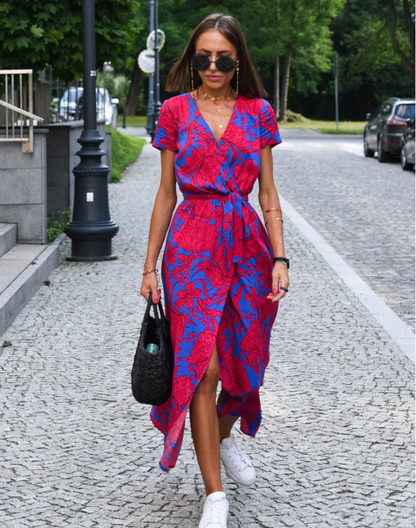 Red - A woman walking on a cobblestone street wearing a vibrant pink and green floral print midi dress with a V-neckline, short sleeves, a tie-waist, and a front slit.